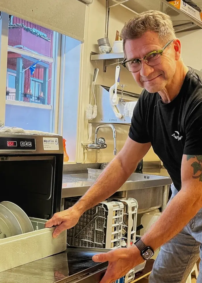 En person lastar ur en diskmaskin i ett kök med stora fönster. Utanför syns röda husväggar. Köket har ljusa väggar och hyllor med köksutrustning.