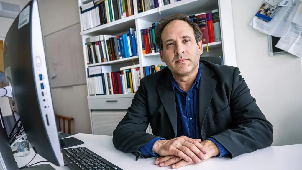 Mark Klamberg, professor i folkrätt vid Stockholms universitet, sitter vid ett skrivbord med en dator och bokhyllor i bakgrunden. Han bär en mörk kavaj och blå skjorta.