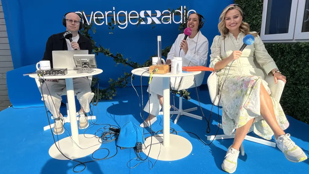 Sveriges Radio sänder utomhus: två programledare vid små bord och en gäst i ljus klänning sitter på blå scen med mikrofoner och kaffekoppar.