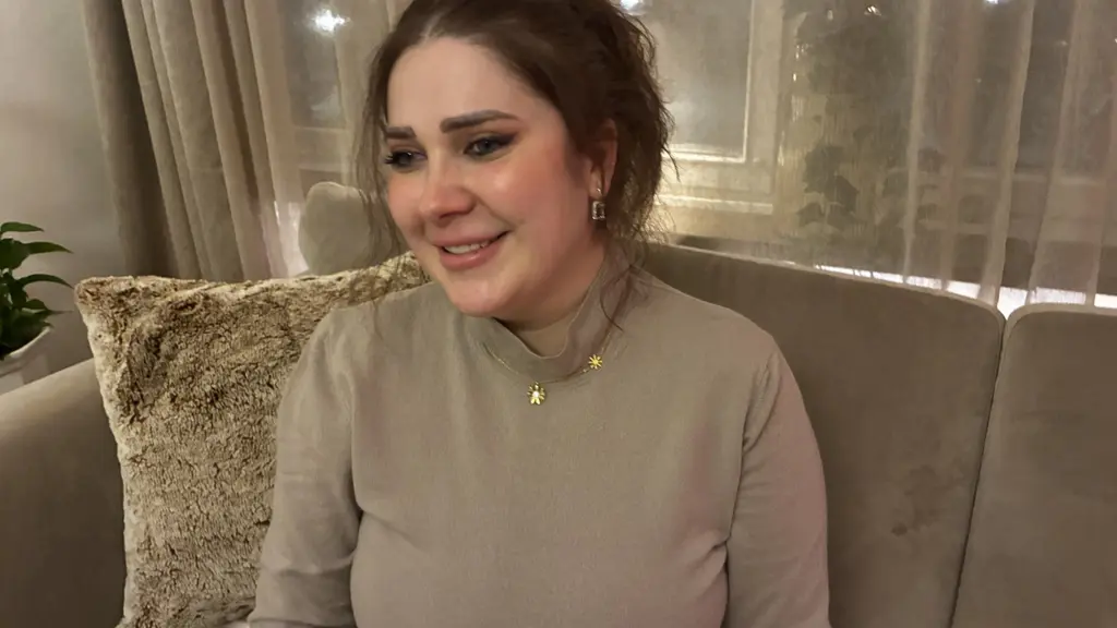 En kvinna sitter på en beige soffa med en ljusgrå tröja. Bakgrunden visar ett fönster med gardiner och svag belysning. En dekorativ kudde syns till vänster på soffan.