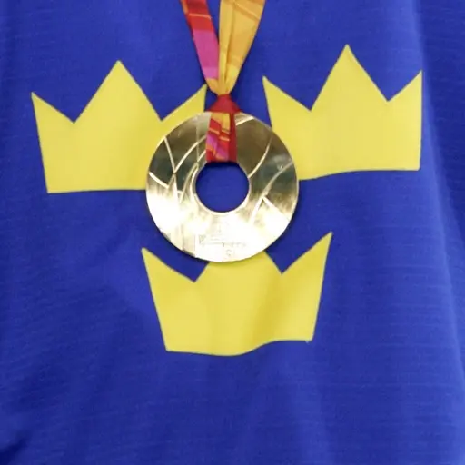 En person klädd i en blå ishockeytröja med Sveriges tre kronor-symbol bär en guldmedalj runt halsen. Medaljen hänger på ett rött band och symboliserar framgång i OS.