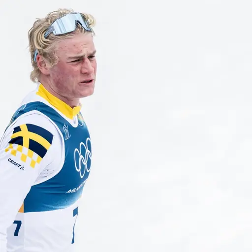 En skidåkare i vit och blå tävlingsdräkt syns på en snötäckt bana. Personen håller stavar och har ett pannband samt skidglasögon på sig.