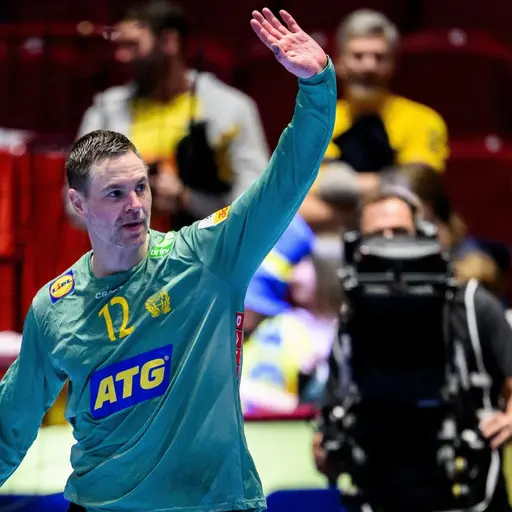 En handbollsspelare i grön målvaktsdräkt med nummer 12 syns på planen, omgiven av publik på läktaren. Spelaren lyfter handen som en gest, med reklam och färgglada detaljer i bakgrunden.