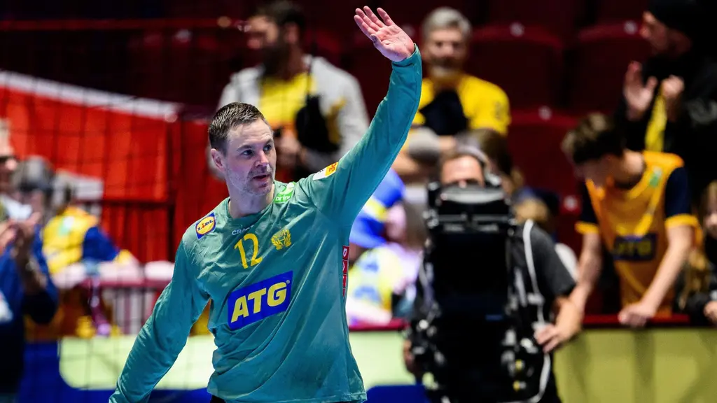 En handbollsspelare i grön målvaktsdräkt med nummer 12 syns på planen, omgiven av publik på läktaren. Spelaren lyfter handen som en gest, med reklam och färgglada detaljer i bakgrunden.