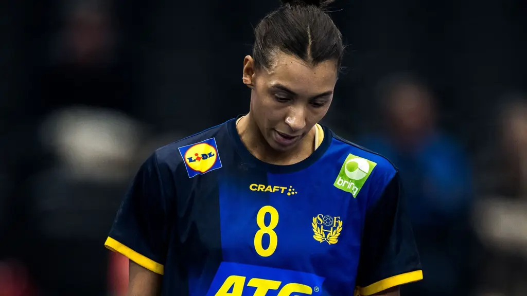 En handbollsspelare i blå och gul matchtröja med nummer 8 syns på planen. Tröjan har sponsorer som ATG och Lidl samt Sveriges emblem. Bakgrunden är mörk med suddiga konturer av publiken.