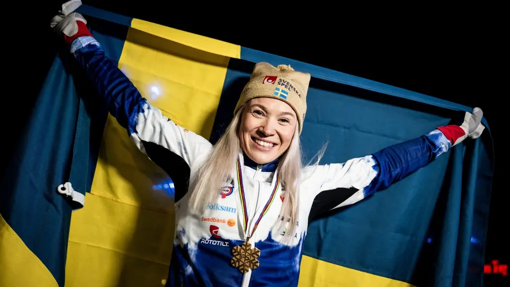 Blond kvinna i gul mössa står och håller i en blågul svensk flagga med guldmedalj runt halsen