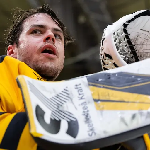 En ishockeymålvakt i gul och svart tröja håller upp sin plockhandske och målvaktsklubba under en match i en arena med takbelysning.