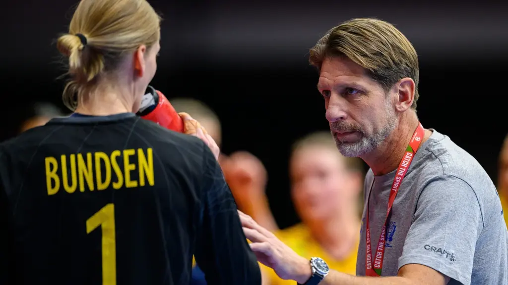 Handbollsspelaren Bundsen med nummer 1 på ryggen samtalar med en tränare under en match. Personen håller en vattenflaska och är klädd i sportutrustning.