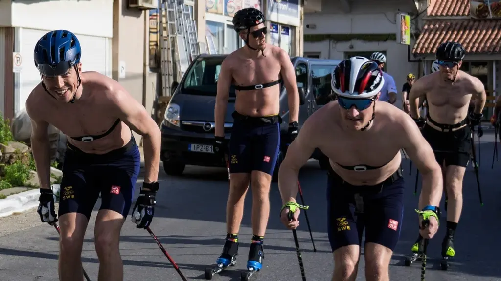 Fyra personer tränar rullskidor på en asfalterad gata i en stadsmiljö. De bär hjälmar och träningskläder, och omgivningen består av bostadshus och parkerade bilar.