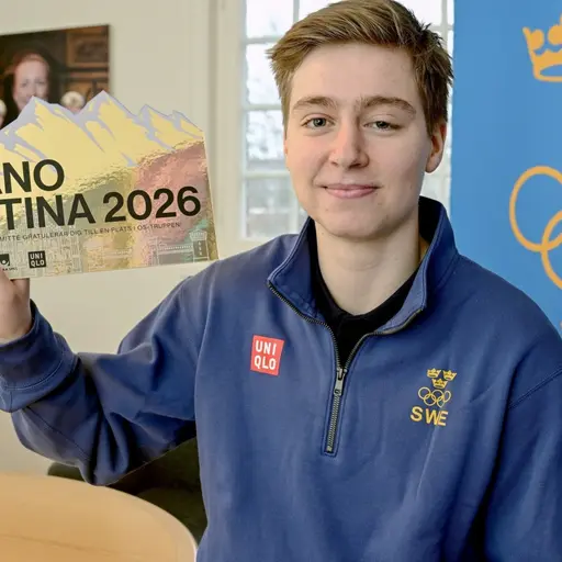 En person håller upp en gul skylt med texten 'Milano Cortina 2026 SWE'. Bakgrunden visar ett fönster, en blå vägg med Sveriges Olympiska Kommittés tre kronor, och en tavla.