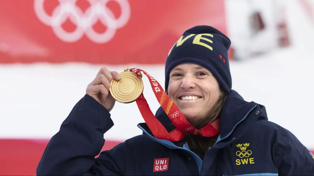 En person klädd i svenska landslagets vinterkläder håller upp en guldmedalj framför en röd bakgrund med OS-ringarna synliga. Bilden är från vinter-OS 2022 vid alpina arenan i Yanqing.