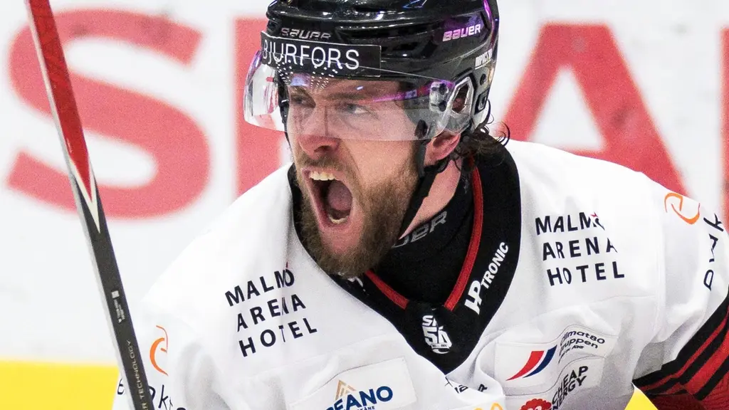 En ishockeyspelare från Malmö IF i vit tröja med texten 'Malmö Arena Hotel' syns på isen under en match mot Timrå. Spelaren håller i en klubba och en reklamvägg är synlig i bakgrunden.