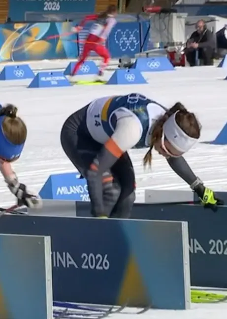 Skidåkare förbereder sig vid startlinjen under en tävling i vinter-OS 2026 i Milano-Cortina. De står böjda över sina stavar på en snötäckt bana med skyltar och OS-logotyper i bakgrunden.