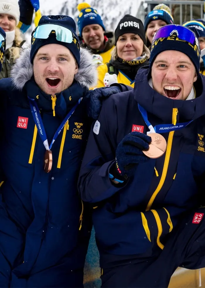 Svenska skidskyttelaget firar en bronsmedalj under ett vinterevenemang. Laget poserar i blåa tävlingskläder med medaljer runt halsen, framför en svensk flagga och glada supportrar.