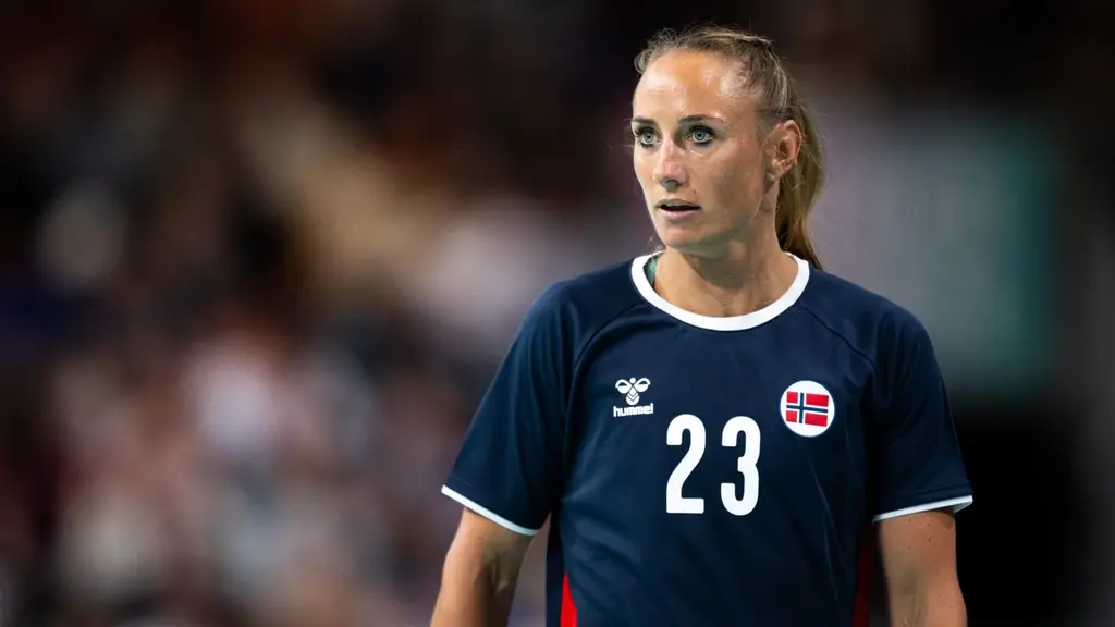 En handbollsspelare från Norge i blå uniform med nummer 23 syns på planen under en match, omgiven av en suddig bakgrund av publik och arena.