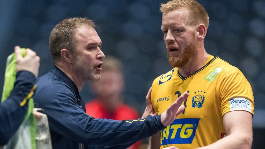 En handbollsspelare i gul matchtröja står på planen och samtalar med en person i mörka kläder. Personen håller en grön handduk. Bakgrunden är en idrottsarena med suddiga detaljer.