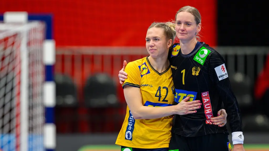 Två svenska handbollsspelare i gula tröjor omfamnar varandra på planen under en match, med en röd och svart bakgrund och målbur i bakgrunden.