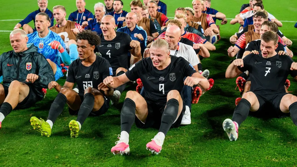 Norges fotbollslandslag ror på planen i segerfirande efter match, med spelare och ledare sittande i rad på gräset framför målet.
