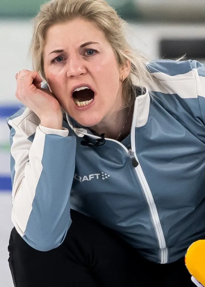 Curlingspelaren Kristin Skanslien hukar sig på isen, iklädd en ljusblå jacka och svarta byxor. Hon håller en gul borste och observerar spelet på en curlingbana med olympiska symboler i bakgrunden.