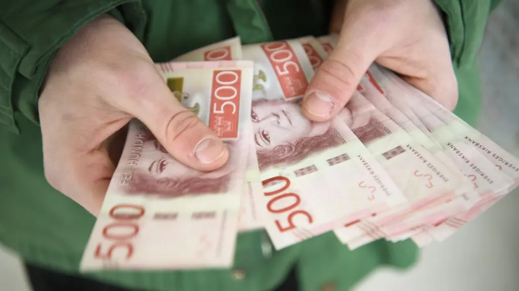 En person håller flera svenska 500-kronorssedlar i händerna. Sedlarna är röda med porträtt av Karl XI, och personen bär en grön jacka. Bilden symboliserar ekonomi och pengar.