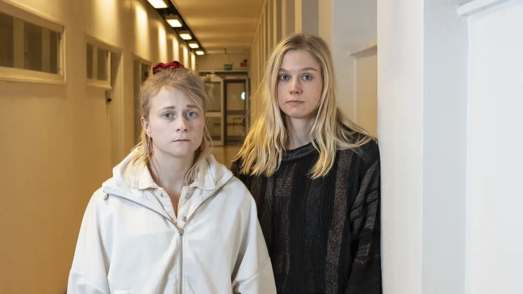 Två kvinnor står i en vit korridor. Den ena har ljusa kläder, den andra mörka. 