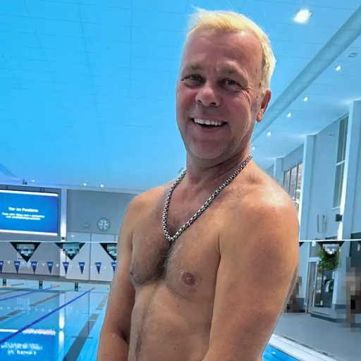 En blond man står barbröstad framför en simbassäng i ett badhus. 