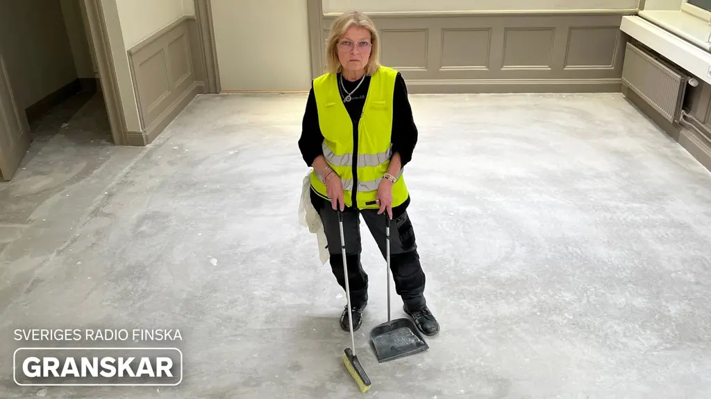 Städaren Tarja Seluska-Kaskilahti iförd gul reflexväst står i ett tomt rum och håller i en mopp. Bilden belyser underrepresentation av personer med finländsk bakgrund inom vissa yrken.