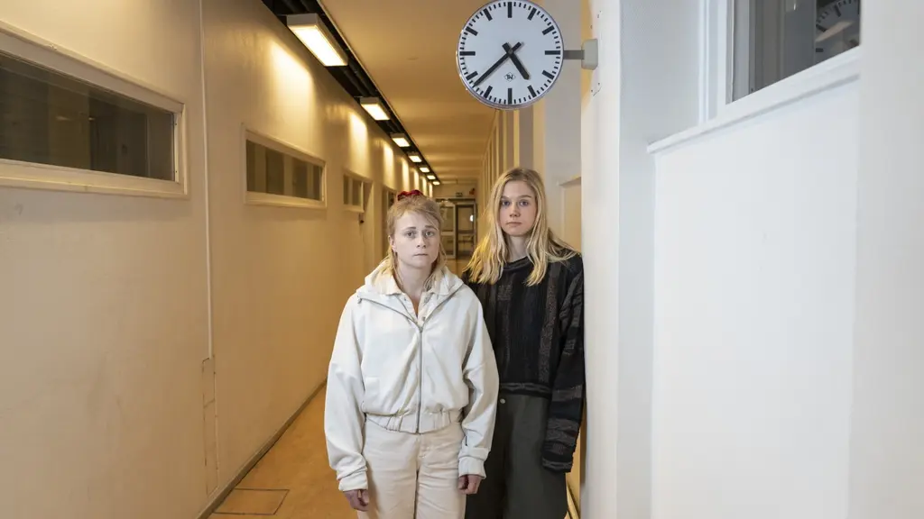 Två kvinnor står i en vit korridor med en väggklocka ovanför dem. Den ena har ljusa kläder, den andra mörka. Korridoren har enkel design med fönster och lysrör i taket.
