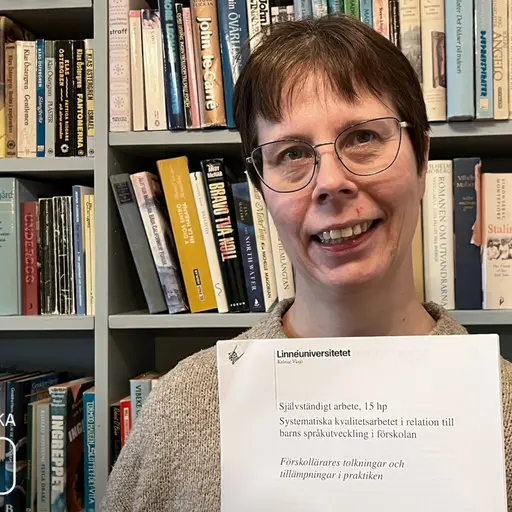 Reija Heikkilä seisoo kirjahyllyn edessä ja pitelee kädessään opinnäytetyötään.