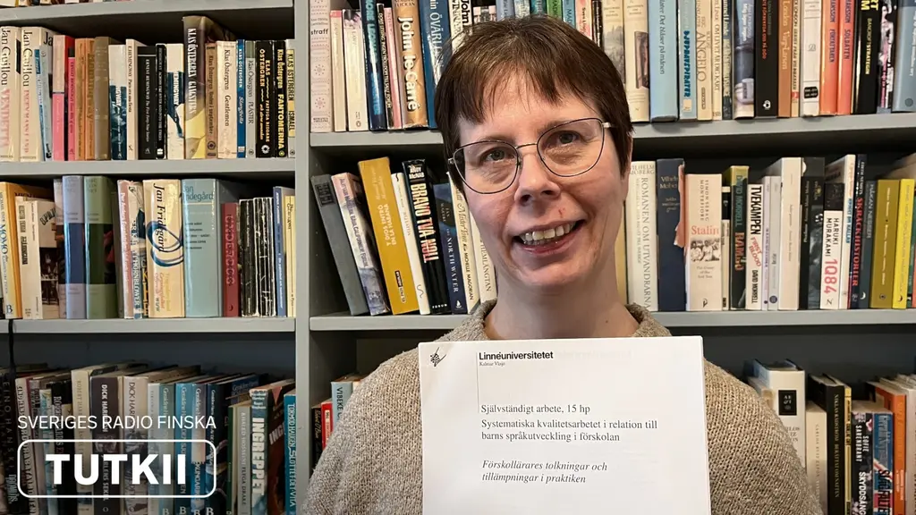 Reija Heikkilä seisoo kirjahyllyn edessä ja pitelee kädessään opinnäytetyötään.