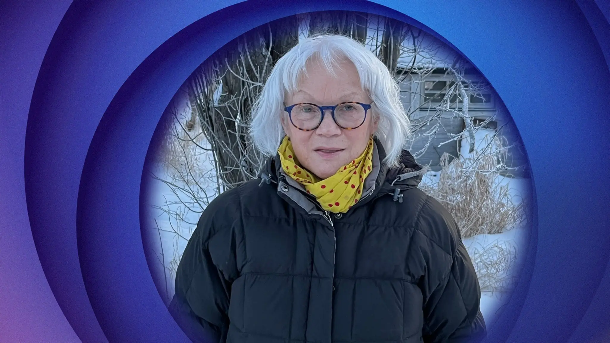 A woman with glasses standing in the snow, wearing a yellow scarf with red polka dots and a black puffer jacket.