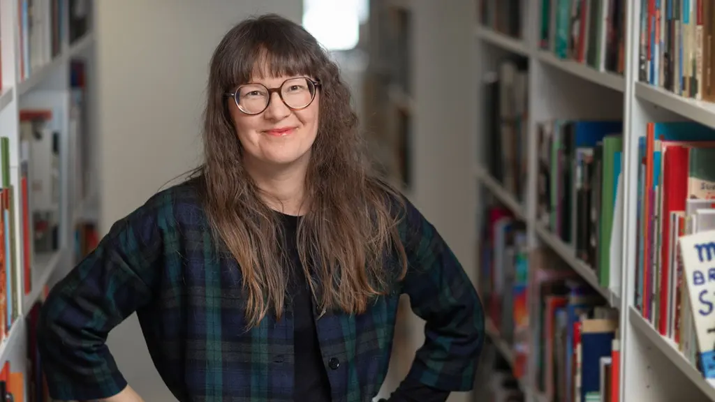 Kvinna ,långt rakt, ganska mörkt hår, glasögon. står leende framför bokhyllor. Hon arbetar på Kulturrådet som handläggare av bidrag 