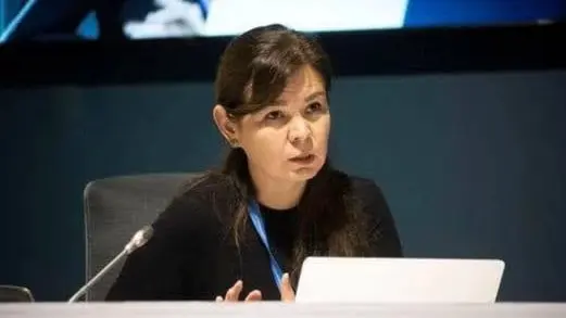 En person sitter vid ett skrivbord utrustat med en mikrofon och en bärbar dator. Bakgrunden visar en mörk skärm med diffusa detaljer. Miljön tycks vara en konferens eller möte.