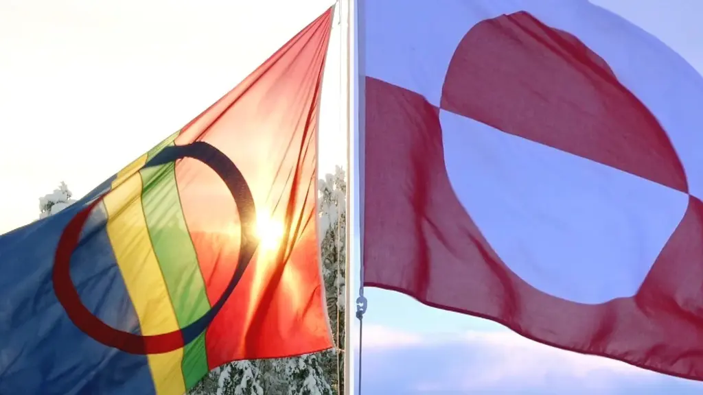 Två flaggor vajar i vinden. Till vänster syns den samiska flaggan med cirkulära färger mot en blå bakgrund, och till höger den grönländska flaggan med röd och vit design. Solen skiner bakom flaggorna.