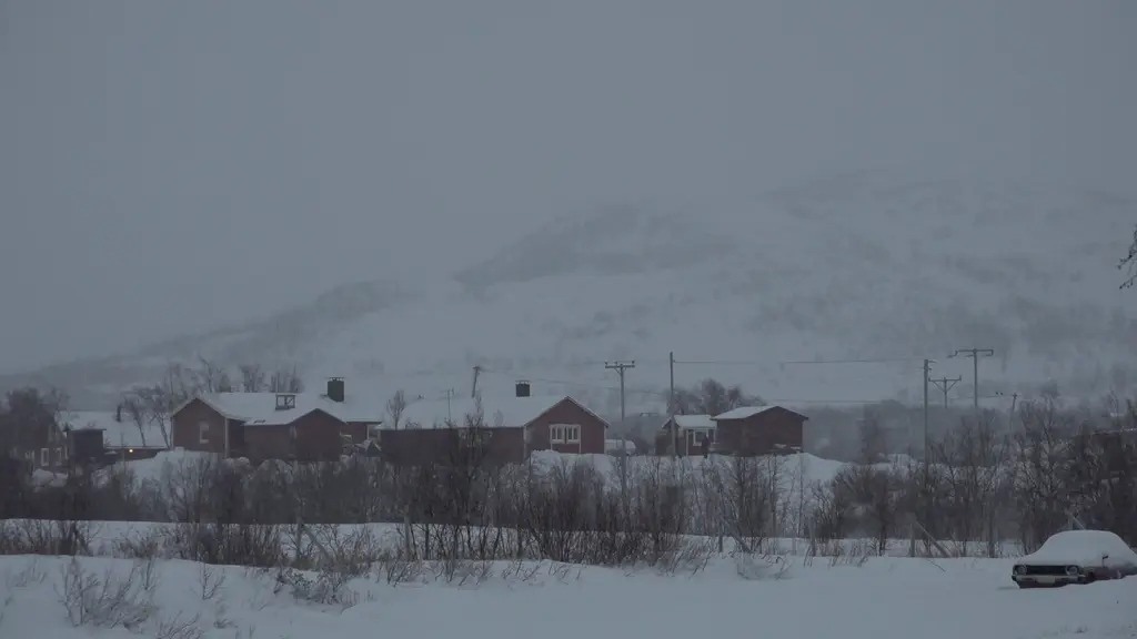 Snötäckt landskap med röda hus som ligger utspridda framför en dimmig bergskedja. Träd och buskar täcker marken, medan en bil står parkerad vid kanten av bilden. Mörk himmel förstärker vinterkänslan.
