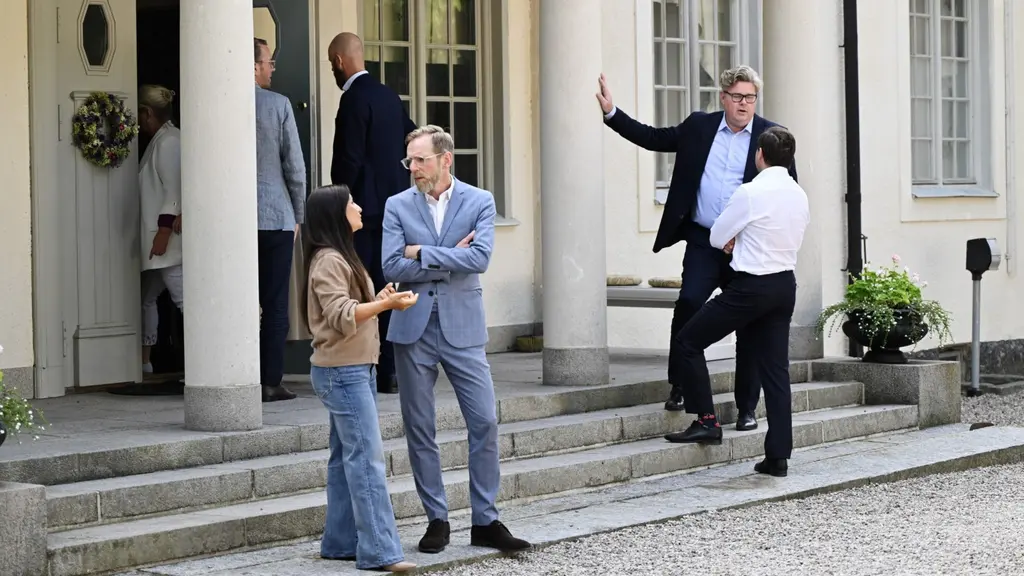 Fyra ministrar står utanför en byggnad vid Harpsund och samtalar. En person gestikulerar medan de andra pratar. Byggnaden har vita pelare och grus framför entrén. Solen lyser svagt.