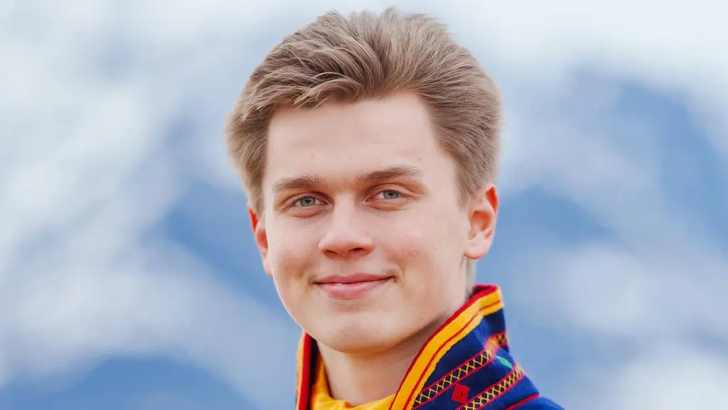 En person klädd i en färgglad dräkt står framför en suddig bakgrund med snötäckta berg och blå himmel. Fokuset ligger på kläderna, som har ett traditionellt mönster och starka färger.