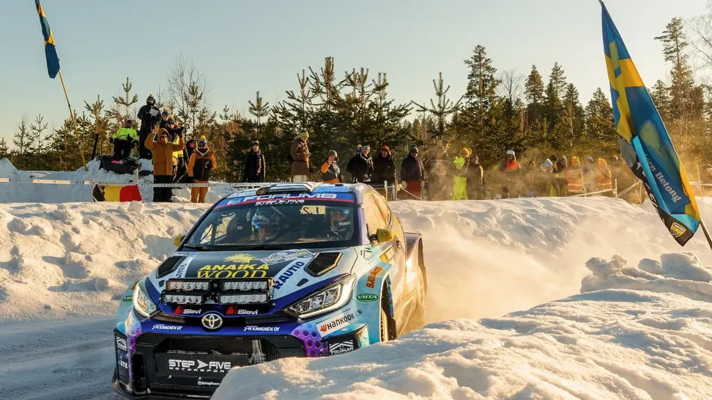 En rallybil, en Toyota GR Yaris, kör snabbt genom en snötäckt sträcka under Svenska rallyt i Umeå. Publik med svenska flaggor syns vid sidan av banan, omgiven av vinterlandskap.