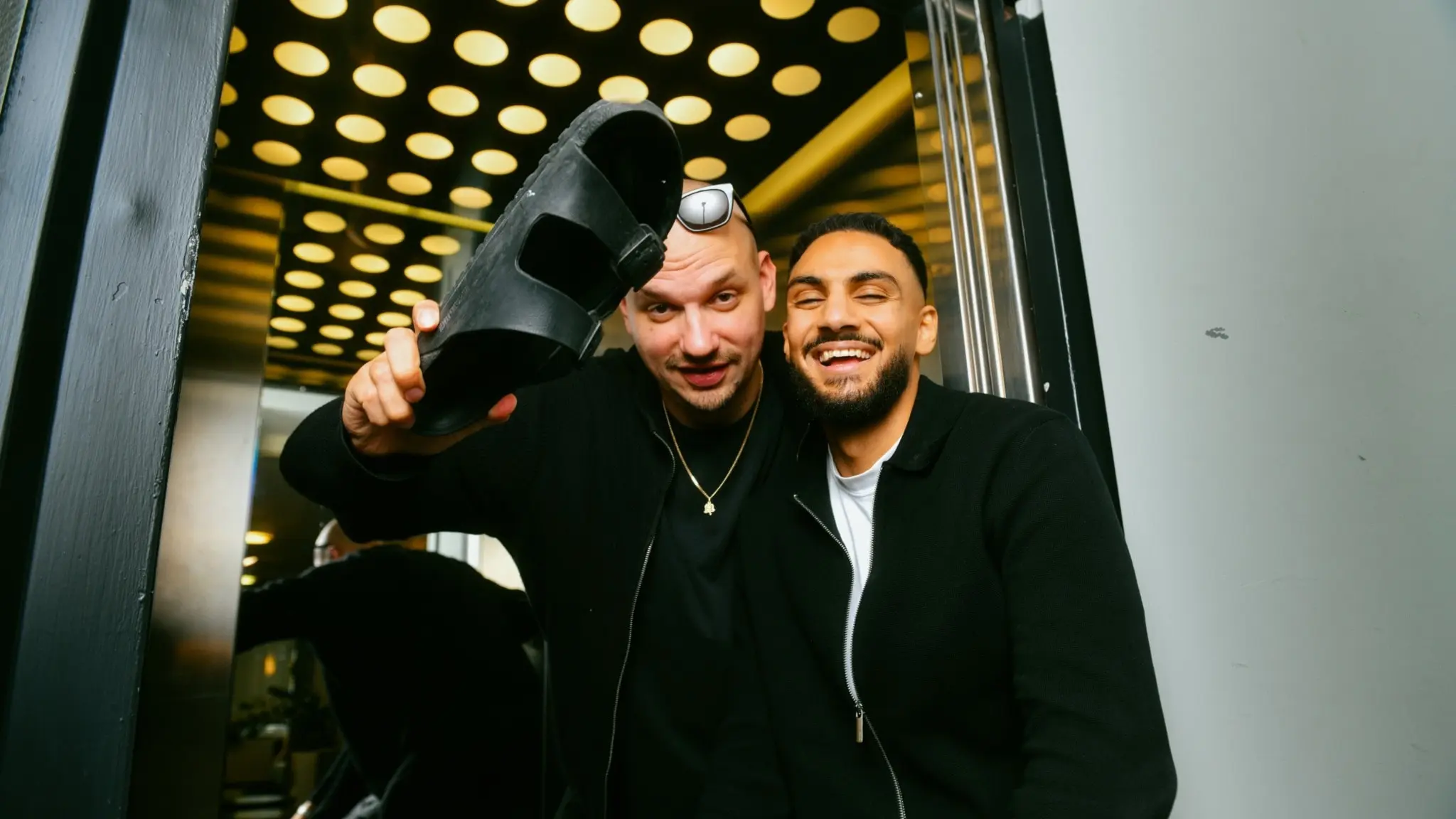 Petter och Haider står vid en hiss. Petter håller en toffla i sin hand.