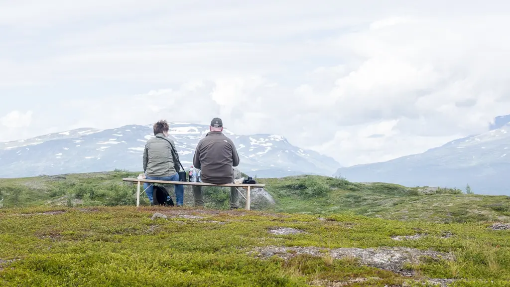 En man och en kvinna sitter på en träbänk uppe på fjället och blickar ut över fjälltoppar, himlen och molnen.