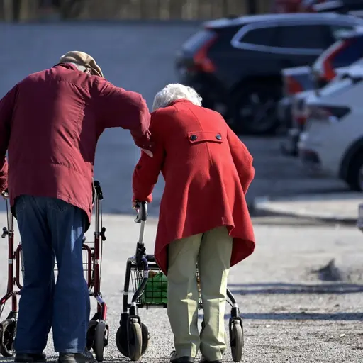 Ett äldre par går med rullatorer på en parkeringsplats i Huvudsta. De är klädda i röda jackor och lutar sig framåt medan de promenerar på den solbelysta platsen.
