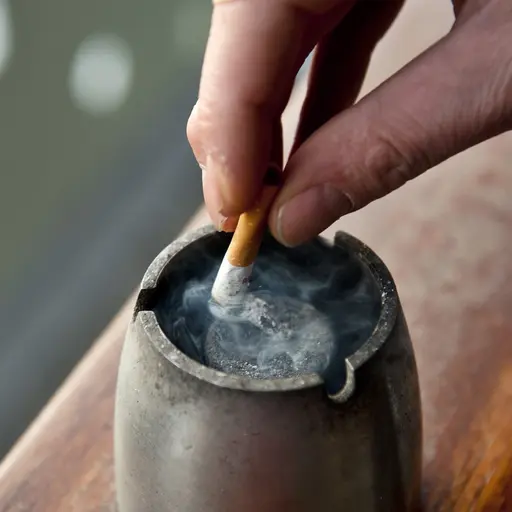 En hand som fimpar en cigarett i ett askfat. Röken från cigaretten stiger uppåt, och askfatet står på en träyta. Bakgrunden är suddig med vita detaljer.