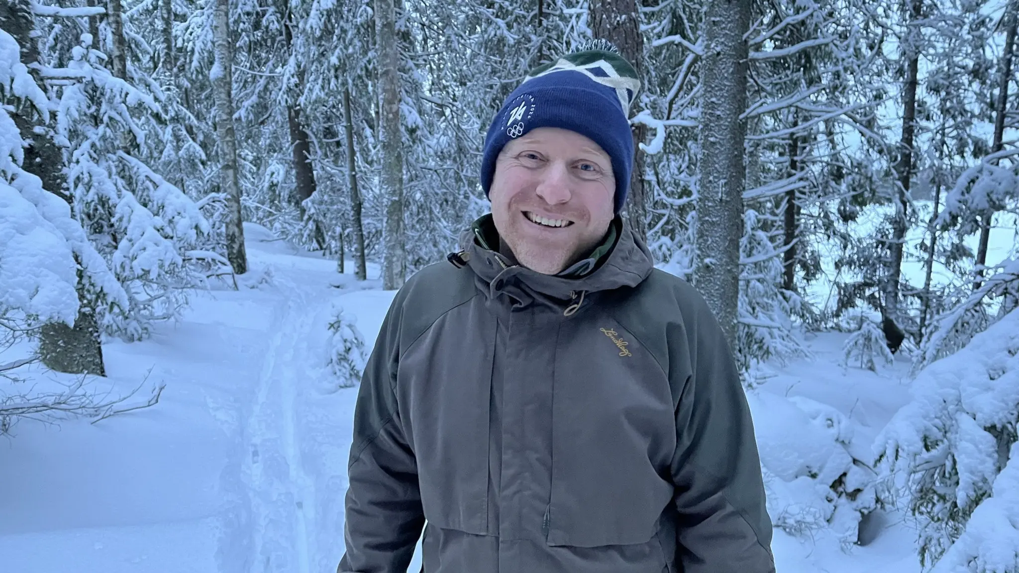 En person klädd i vinterjacka och mössa står i en snöig skog i Gästrikland. Träden är täckta av snö och skidspår i bakgrunden.