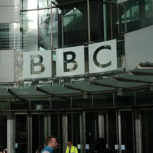 En person går förbi BBC:s huvudkontor i London, en byggnad med glasfasad och tydlig BBC-skylt ovanför ingången. Reflektioner av ljus syns på glaset, och en säkerhetsvakt står vid entrén.