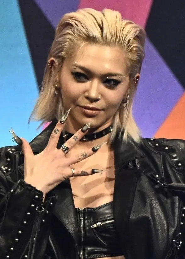 En person i svart läderklädsel står framför en färgglad logotyp för Melodifestivalen 2026. Personen poserar självsäkert med handen vid halsen och bär en kort topp och jacka.