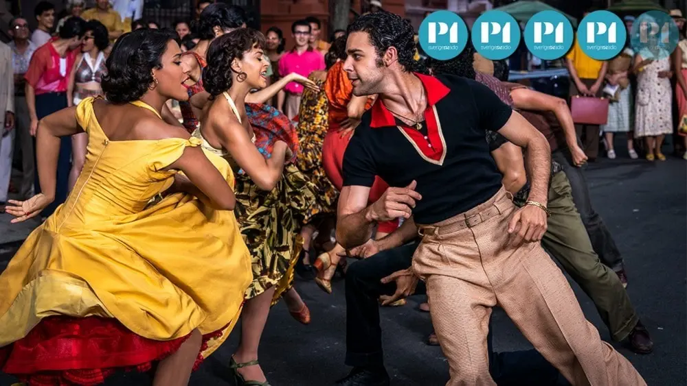 Stillbild ur filmen West side story av Steven Spielberg. Ariana DeBose som Anita, klädd i gul klänning, och David Alvarez som Bernardo, klädd i beiga byxor och kortärmad svart tröja med röd krage, dansar med varandra.