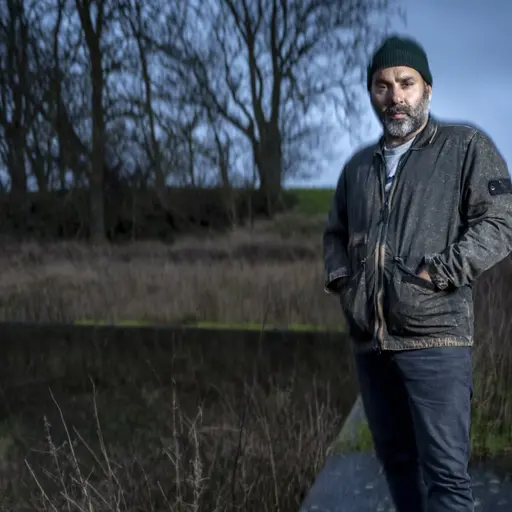 En man står utanför ett hus. Gräs och träd i bakgrunden. Lantligt.