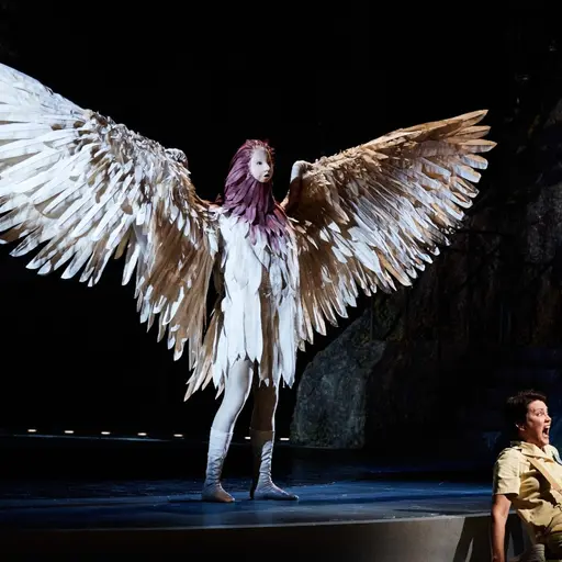 En scen från en operaproduktion där en person med stora, vita änglavingar står upplyst på scenen, medan en annan person sitter på golvet i en dramatisk position. Bakgrunden är mörk och mystisk.