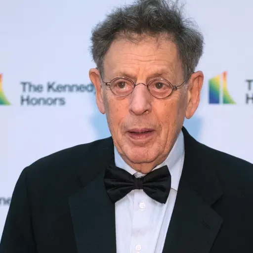 En person klädd i smoking med fluga står framför en vit bakgrund med texten 'The Kennedy Center Honors' och en färgglad logotyp. Bilden är tagen vid ett formellt evenemang.