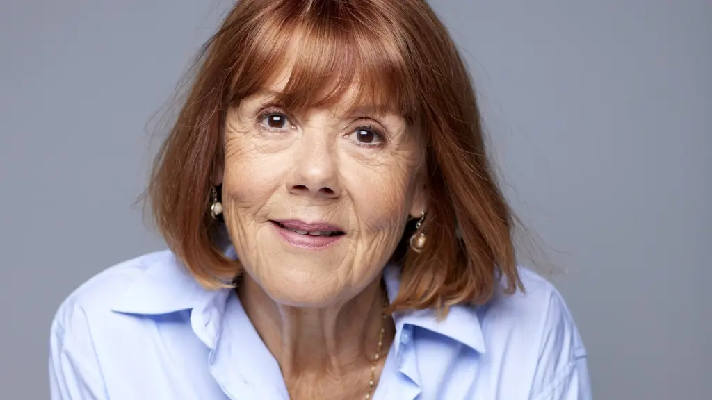 En person med axellångt rött hår bär en ljusblå skjorta och ett halsband. Bakgrunden är neutral med grå ton